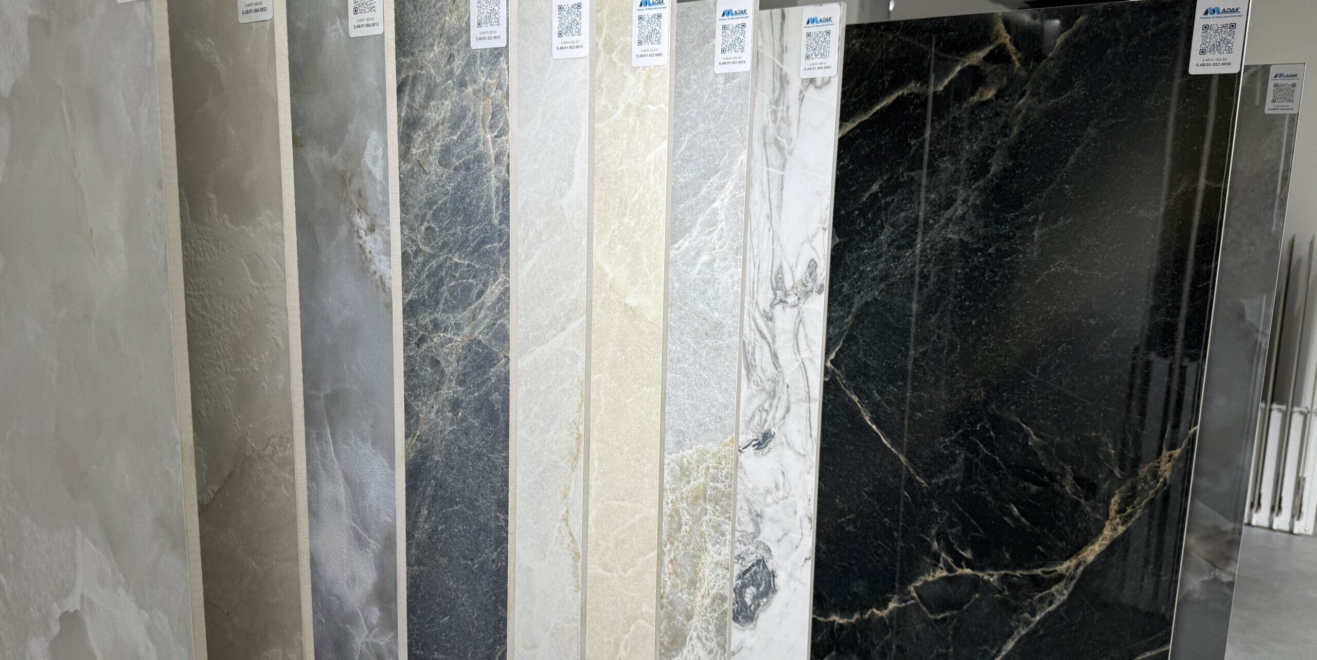 Große Fliesenauswahl bei Madak Mannheim – Natursteinoptik Fliesen in 80x160 Format, matt und poliert, mit schwarzer polierter Fliese und goldenen Marmorierungen im Vordergrund.